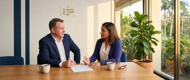 A senior delivery lead in a navy blazer and a prospective client in an indigo cardigan seated across a long warm-timber table at late afternoon, mid-conversation, only a single set of handwritten notes between them with a fine-liner pen poised over the page, two mugs of tea and a folded pair of reading glasses beside, a small cream-paper card on the wall behind reading the function the standard the gap, fiddle-liner pen across the top folder, fiddle-leaf fig nearby, subtropical Gold Coast greenery visible through floor-to-ceiling glass, warm afternoon light from the right.