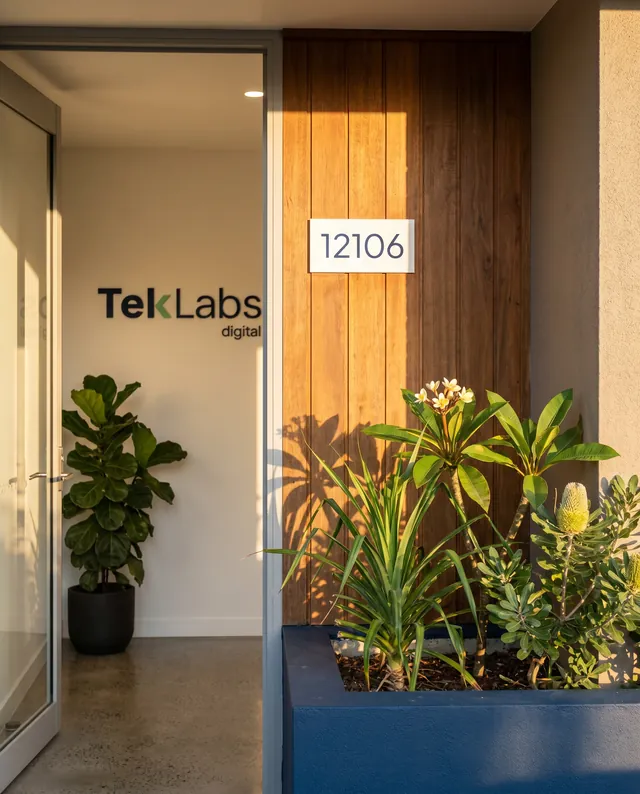 A quiet exterior corner of the Gold Coast headquarters at 12106 The Darling Ave, Broadbeach, at late afternoon, warm-timber-and-render wall meeting a low painted-navy planter with a young pandanus, a small frangipani in bloom, and a thriving banksia, a small paper-white house number plate mounted at eye-height on the timber reading 12106, a glass studio door slightly ajar with a fiddle-leaf fig visible inside and the full Teklabs Digital lockup visible on the interior wall through the open door, warm afternoon light from the right.