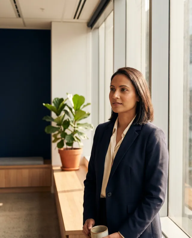 A composed mid-30s Australian professional in a navy blazer over a cream blouse, three-quarter profile near a floor-to-ceiling window in a contemporary Sydney workplace at late afternoon, mid-thought, holding a ceramic mug of tea, a fiddle-leaf fig in a terracotta pot and a deep-navy feature wall in soft focus behind, warm afternoon light from the right.